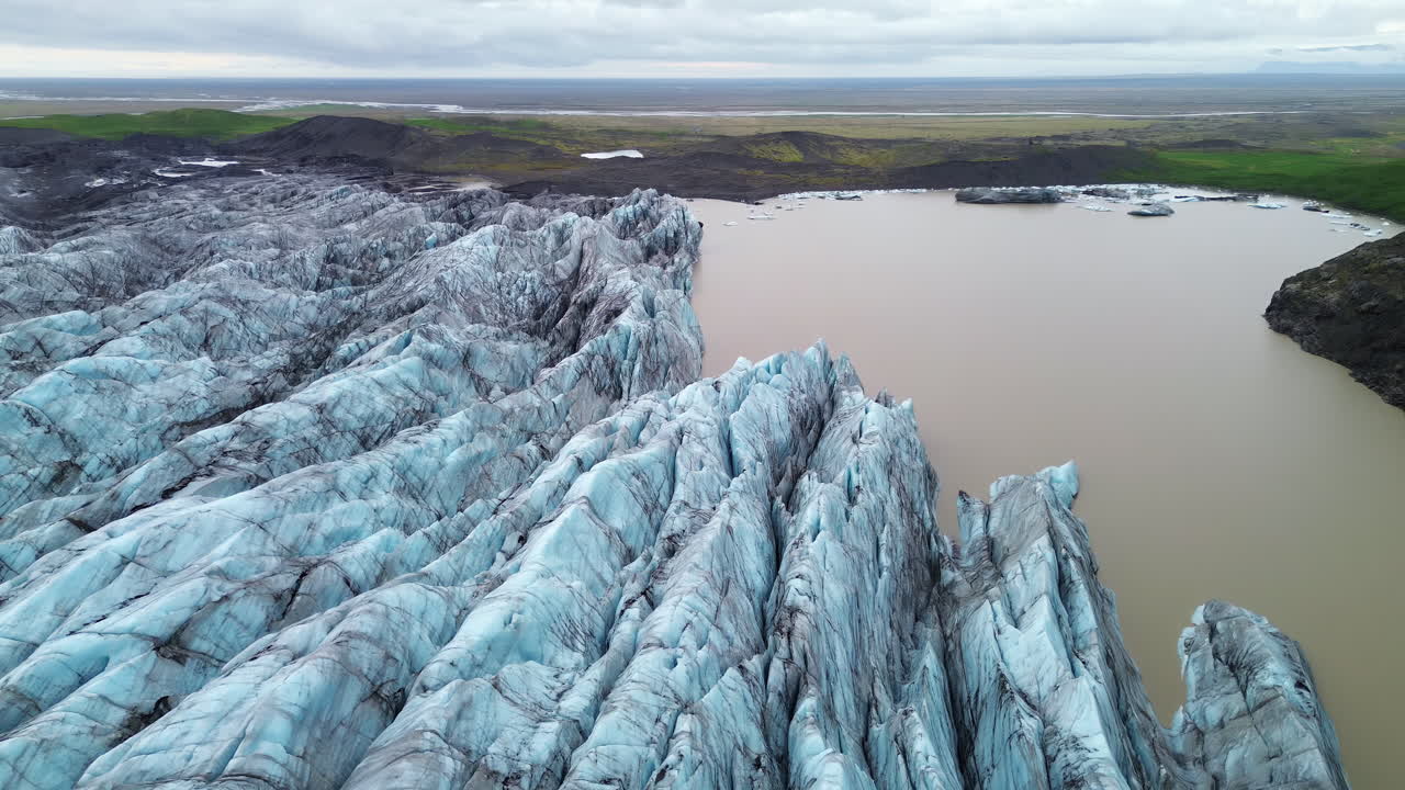 Dronebillede fra luften af Falljökull-gletsjerens dybe sprækker, en udløber af Vatnajökull i Sydisland. Dramatiske blå isrygge og mørke vulkanske askestriber skaber et storslået arktisk landskab under en overskyet himmel.