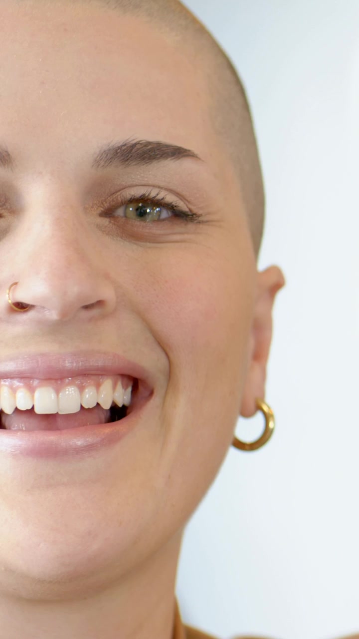 Video verticale: Donna rasata sorridente che allarga la bocca in una risata in studio, indossando un top color cuoio e un anello al naso.