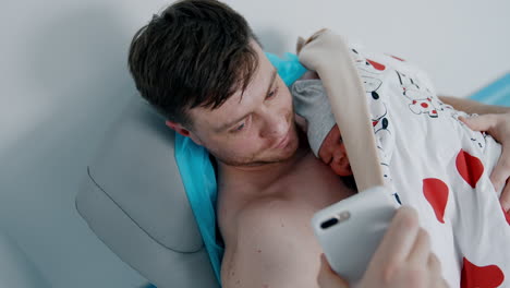 caucasian father is taking video on the phone. happy dad showing his newborn baby lying on his chest. top view.