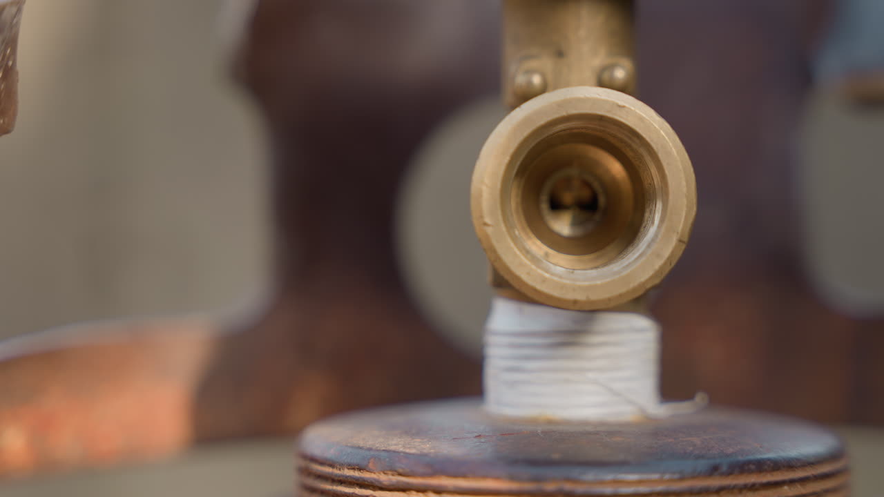 Primo piano di una valvola in ottone su un fornello, scena di manutenzione domestica che mostra un ugello filettato avvolto con nastro bianco, sfondo di metallo arrugginito, profondità di campo ridotta, la calda luce del sole che evidenzia la texture.