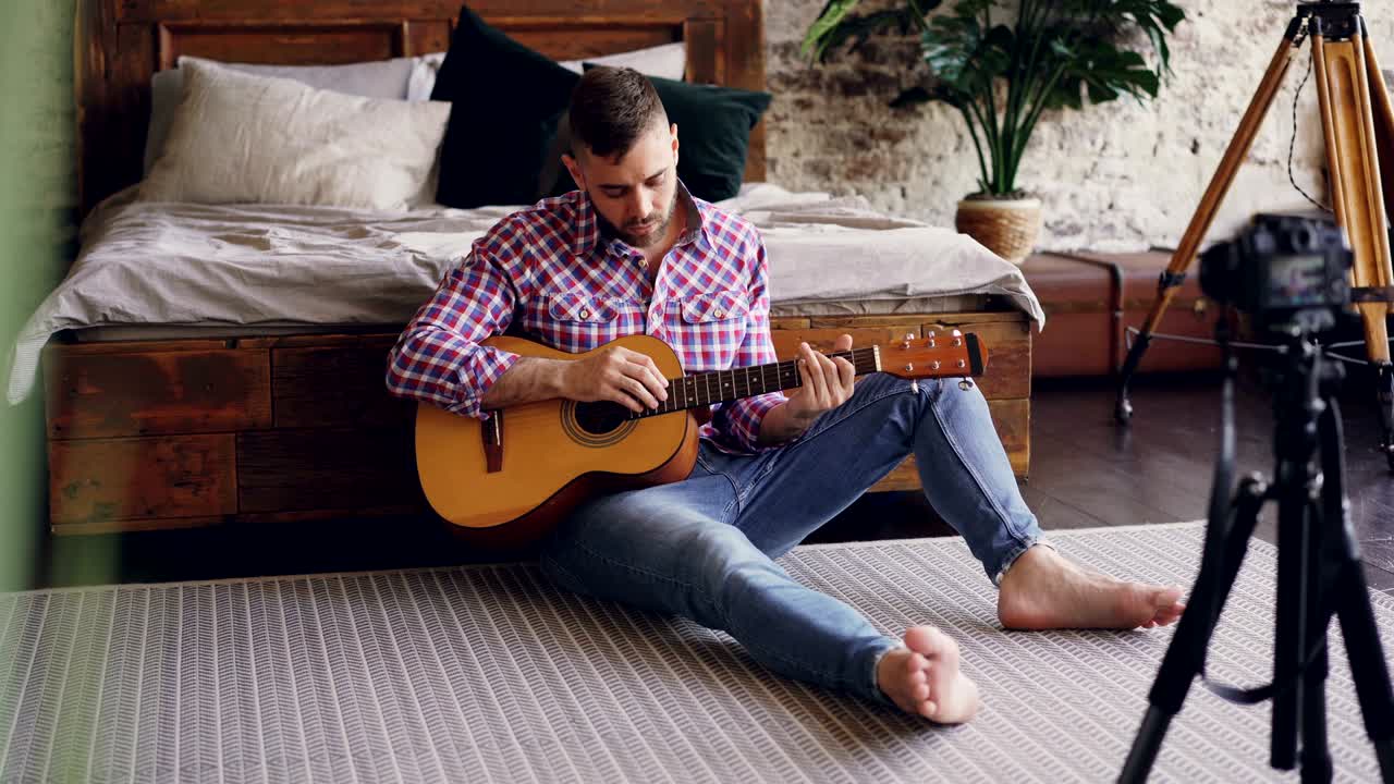 Handsome man blogger and guitarist is recording video lesson about guitar using camera. Bearded guy is greeting his followers, talking and showing guitar parts.