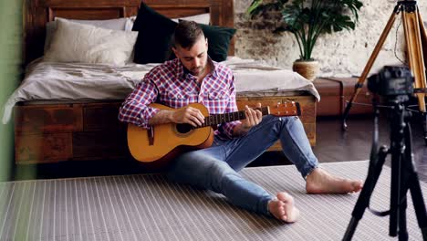 handsome man blogger and guitarist is recording video lesson about guitar using camera. bearded guy is greeting his followers, talking and showing guitar parts.