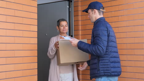 Mujer-De-Pie-En-La-Puerta-Y-Recibiendo-El-Paquete-Del-Servicio-De-Mensajería-Y-Firmando-El-Portapapeles