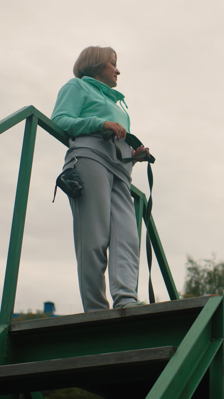 Mujer caucásica en lo alto de unas escaleras verdes en primer plano a ángulo bajo, sujetando una correa con una mano y una bolsa de cámara en la cadera, con la mirada de perfil hacia el horizonte arbolado bajo un cielo cubierto con una sudadera con capucha color menta, gris