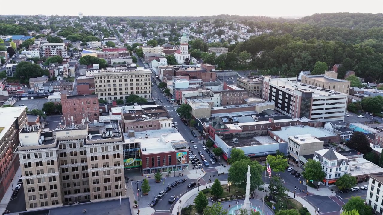 Historisch stadsplein vormt het hart van downtown Easton, Pennsylvania, met een hoog stenen monument en Amerikaanse vlag die oprijzen nabij een ronde fontein te midden van boomrijke straten bij gouden zonsondergang. Brede luchtopname met drone