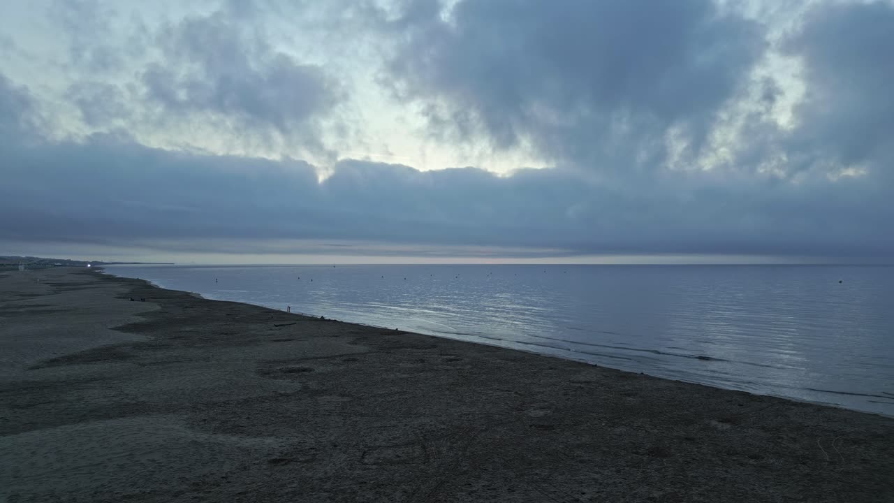 Serene sunrise illuminates a deserted beach, casting soft light over the quiet sea. Clouds drift lazily across the sky, creating a tranquil atmosphere on this idyllic shoreline