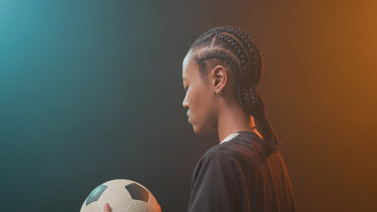 Donna afroamericana in tenuta sportiva che sistema il pallone da calcio, di fronte alla telecamera con luci verde acqua o arancioni, spazio per testo
