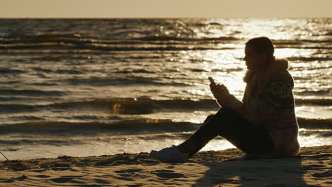 Young-Woman-Sitting-On-The-Beach-At-Sunset-Enjoy-The-Phone-Cool-Autumn-Day