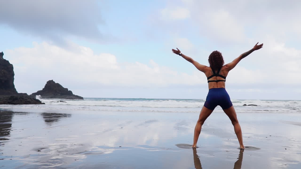 In slow motion, a young, slim woman practices squats and jumps, working out on the ocean's shore