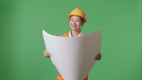 asian female engineer with safety helmet looking at blueprint in her hands while standing in the green screen background studio