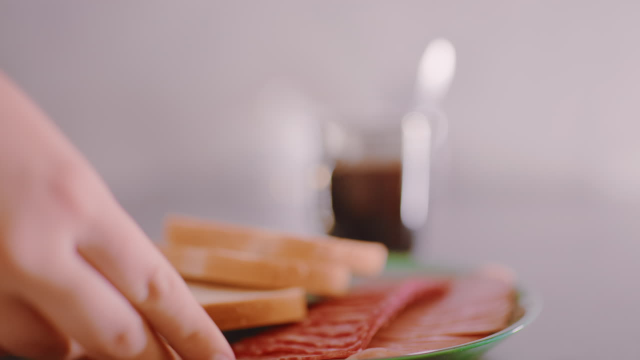 Close up of sliced bread and meat on green plate placed on table with blurred background showing hot coffee cup, emphasizing detail of simple homemade meal in warm indoor kitchen setting