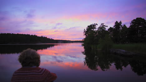 disparo ascendente sobre un hombre sentado en una orilla rocosa, de un lago, un cielo púrpura, en una colorida puesta de sol o atardecer, en albysjon, tyreso, suecia