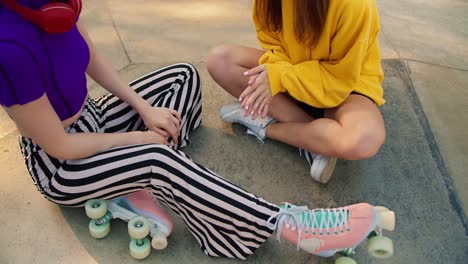 Close-up-shot-of-a-girl-in-a-yellow-sweater-and-a-girl-in-a-purple-top-and-striped-pants-in-pink-roller-skates-sit-on-a-concrete-floor-in-a-skate-park-in-summer