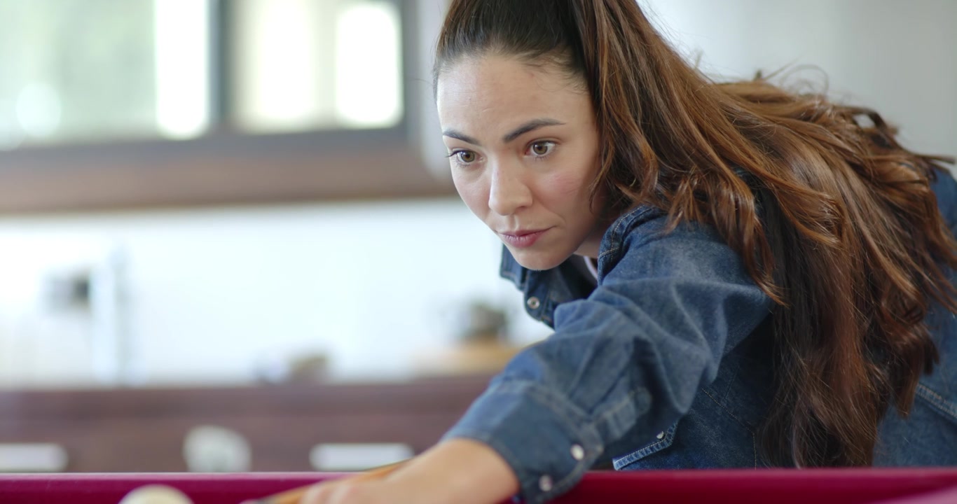 Adult woman aiming to pocket ball, leaning on red-felt pool table, aligning cue, striking cue ball