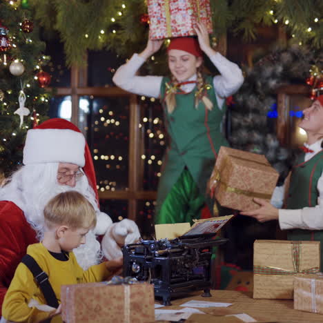 happy elves playing with boxes near santa. santa is typing a letter on old machine near the christmas tree. joyful atmosphere at christmas.