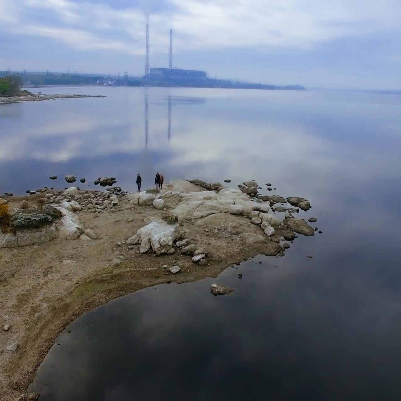 Smooth surface of river on the grey windless autumn day. People standing on the rocky bank of the river. Silhouette of the industrial plant at backdrop