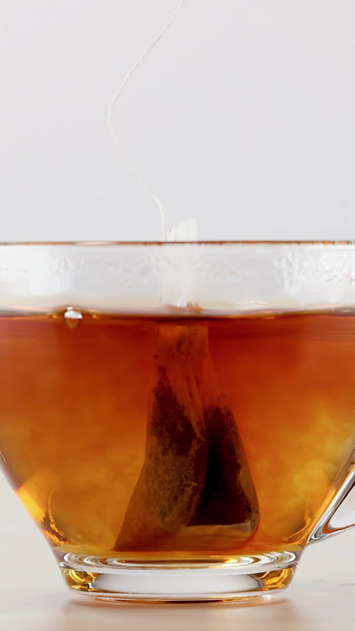 Primo piano di una bustina di tè in infusione in una tazza trasparente con un infuso di erbe che turbina.