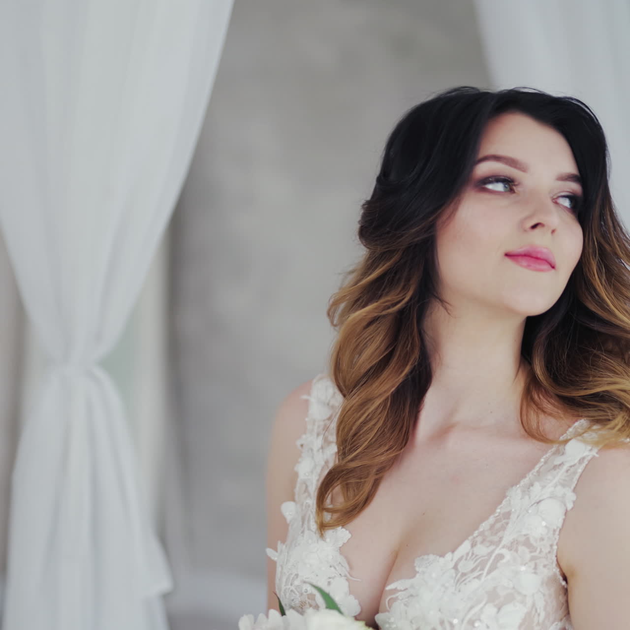 Beautiful young model in a white lace dress sits on a white bed. Gorgeous bride in wedding dress with flowers is sitting in the light bedroom.