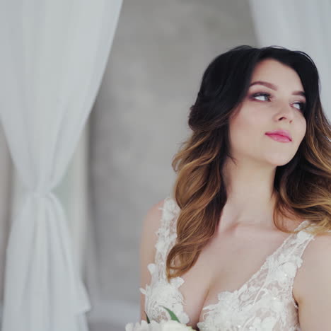 beautiful young model in a white lace dress sits on a white bed. gorgeous bride in wedding dress with flowers is sitting in the light bedroom.