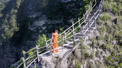 woman admiring diamond beach in nusa penida island indonesia from above the cliffside staircase, aerial dolly out reveal shot