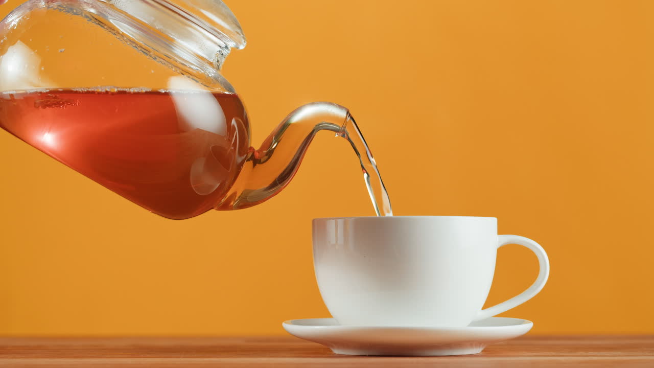 Hot tea being poured from a glass teapot into a white cup