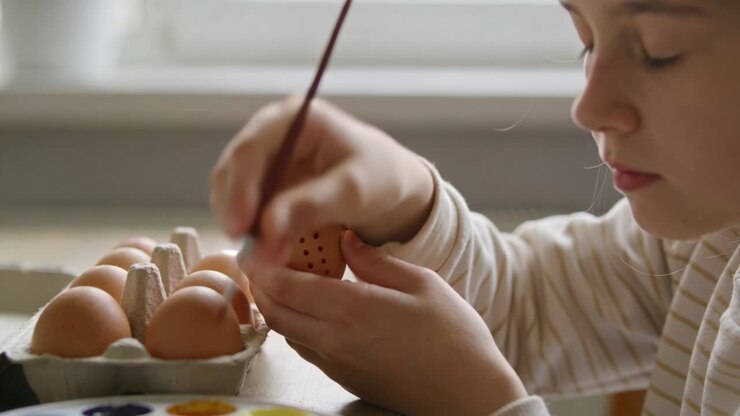Painting girl at table by window, applying dot eggshell design with brush, striped top, copy space