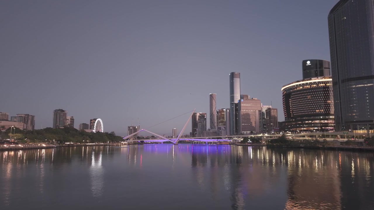 An aerial view at dusk shows the illuminated Brisbane skyline and a purple lit bridge reflected in the calm river.