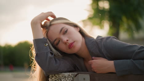 woman resting her head on arm while seated on bench, gently touching her hair as she drops the hand, soft sunlight creates a warm, peaceful ambience around her, with blurred background