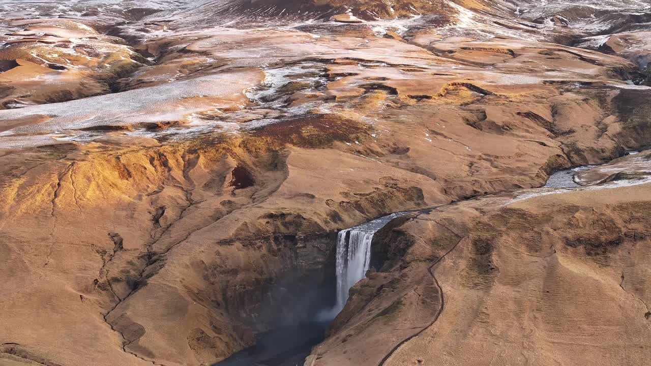 Wide aerial view of Skógafoss waterfall flowing through golden and snow-dusted Icelandic terrain, framed by rugged highlands near Eyjafjallajökull volcano.