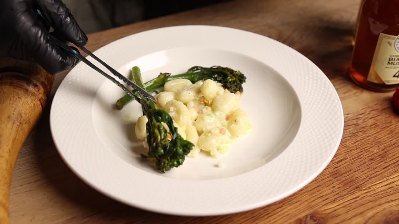 Chef che impiattano gnocchi con salsa cremosa e broccoli su un piatto bianco.