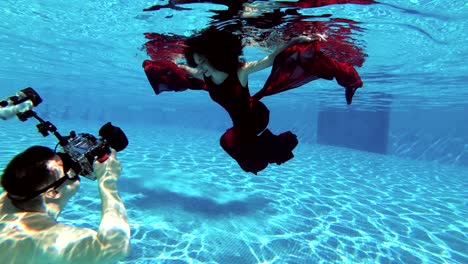 a man photographer takes a professional underwater camera beautiful girl bride with red hair, who swims underwater in the pool in a red dress. slow motion.