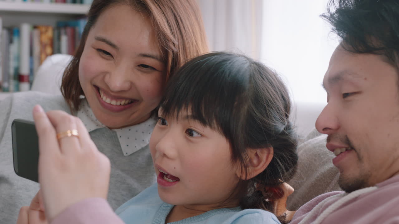 행복한 아시아 가족이 집에서 스마트폰을 사용하여 비디오 채팅을 하고 있습니다. 어머니와 아버지는 딸과 함께 휴대전화로 채팅하고 손을 흔들며 온라인 커뮤니케이션 4k 영상을 즐기고 있습니다.