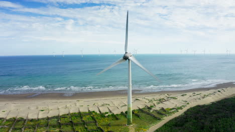 aerial view of rotating wind turbine on sandy beach and wind farm in background standing in water of ocean