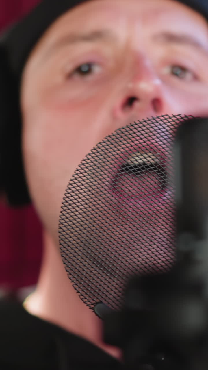 A man dressed in black is wearing headphones and standing in front of a microphone in a red soundproofed room. He is singing with seriousness, making a hand gesture while focusing on his performance