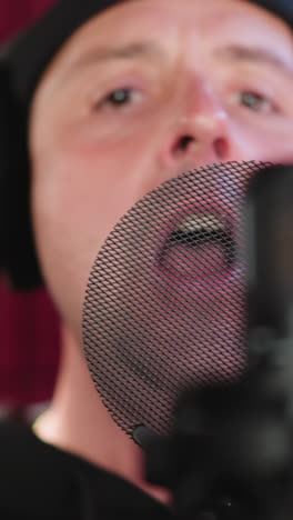 un hombre vestido de negro está usando auriculares y de pie frente a un micrófono en una habitación a prueba de sonido roja. está cantando con seriedad, haciendo un gesto de la mano mientras se centra en su actuación