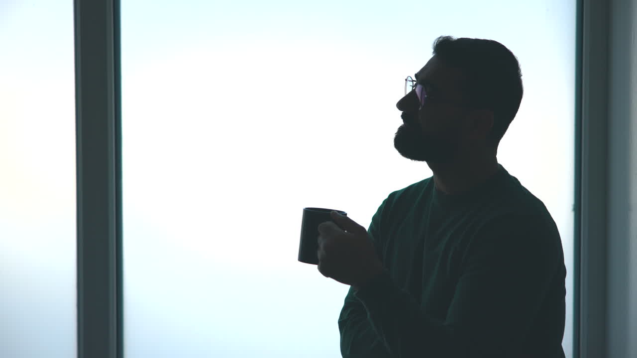 Silhuet af mand drikker kaffe ved vinduet, fjernudvikler holder en stille morgenpause, mens han kigger ud. Medie