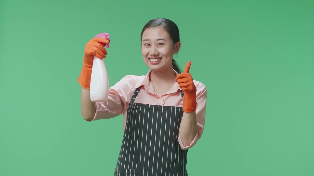 손에 스프레이를 들고 있는 아시안 여성 집주인이 그린 스크린 스튜디오에서 카메라를 향해 엄지손가락을 올리고 있다.