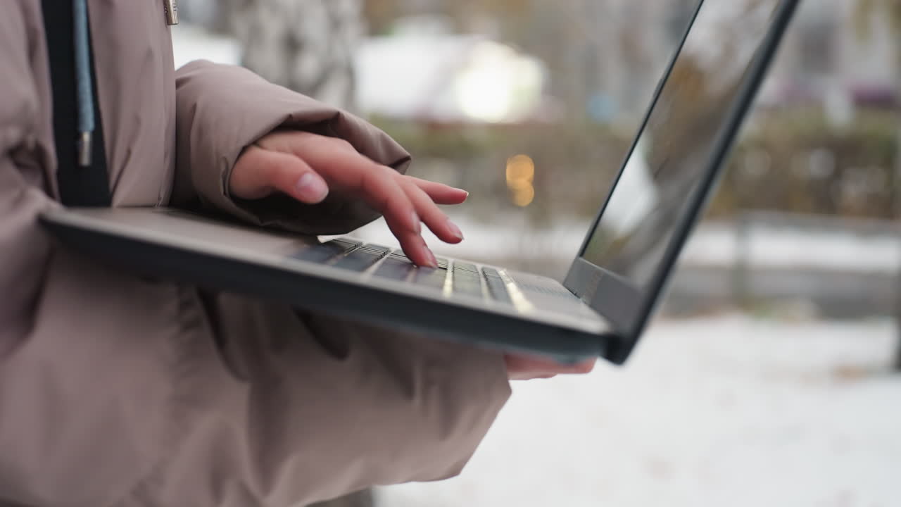 Close-up of light-skinned female hands holding and operating black laptop outdoors in snowy setting, with blurred screen and soft bokeh light in background, fingers actively typing in cold weather