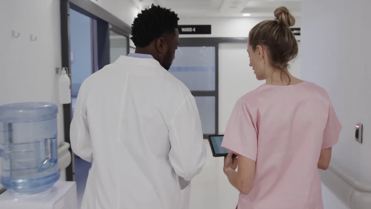 Diverse female nurse and male doctor walking in corridor, using tablet and talking, slow motion