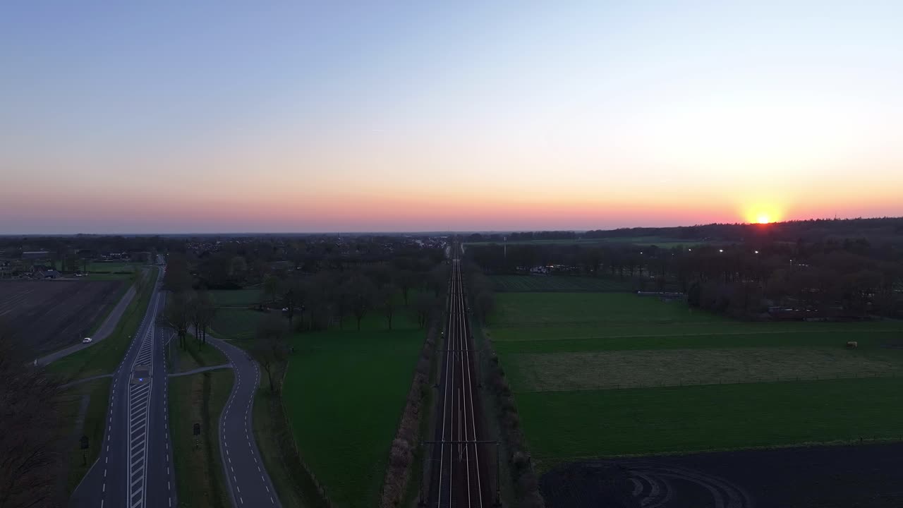 Parallel railway tracks and a curving country road at sunrise, fields and scattered trees on both sides, horizon glowing with early morning light, drone pull out, real time