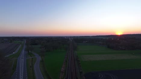 parallel railway tracks and a curving country road at sunrise, fields and scattered trees on both sides, horizon glowing with early morning light, drone pull out, real time