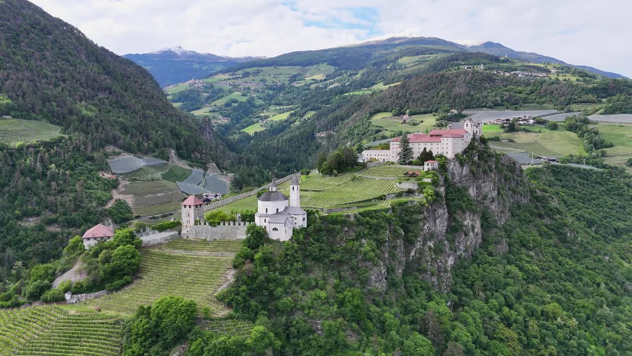 Monastero di Sabio on a hilltop overlooks a lush valley in South Tyrol, with green fields, forested hills, and distant snow-capped mountains