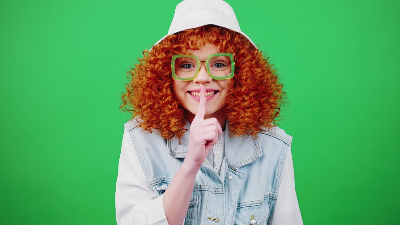 Secret concept. Young mysterious redhead woman looking around and putting finger on lips, green chroma key background