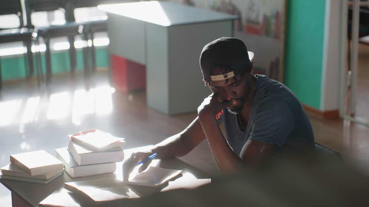 Reflective man sits in library with pen in hand, surrounded by stacked books, lost in deep thought as sunlight streams through windows, creating warm atmosphere of focus and quiet concentration