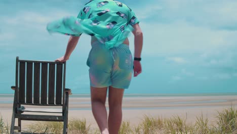 plano general de un joven sentado en una sola silla de playa de madera mirando hacia una playa soleada en marea baja con vetas de agua que se extienden hacia donde el cielo está creando una tormenta furiosa