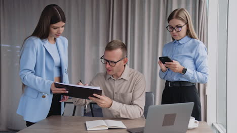 deadline time in the modern office. busy male manager sitting at desk and two female employees standing beside him. man signing the documents.
