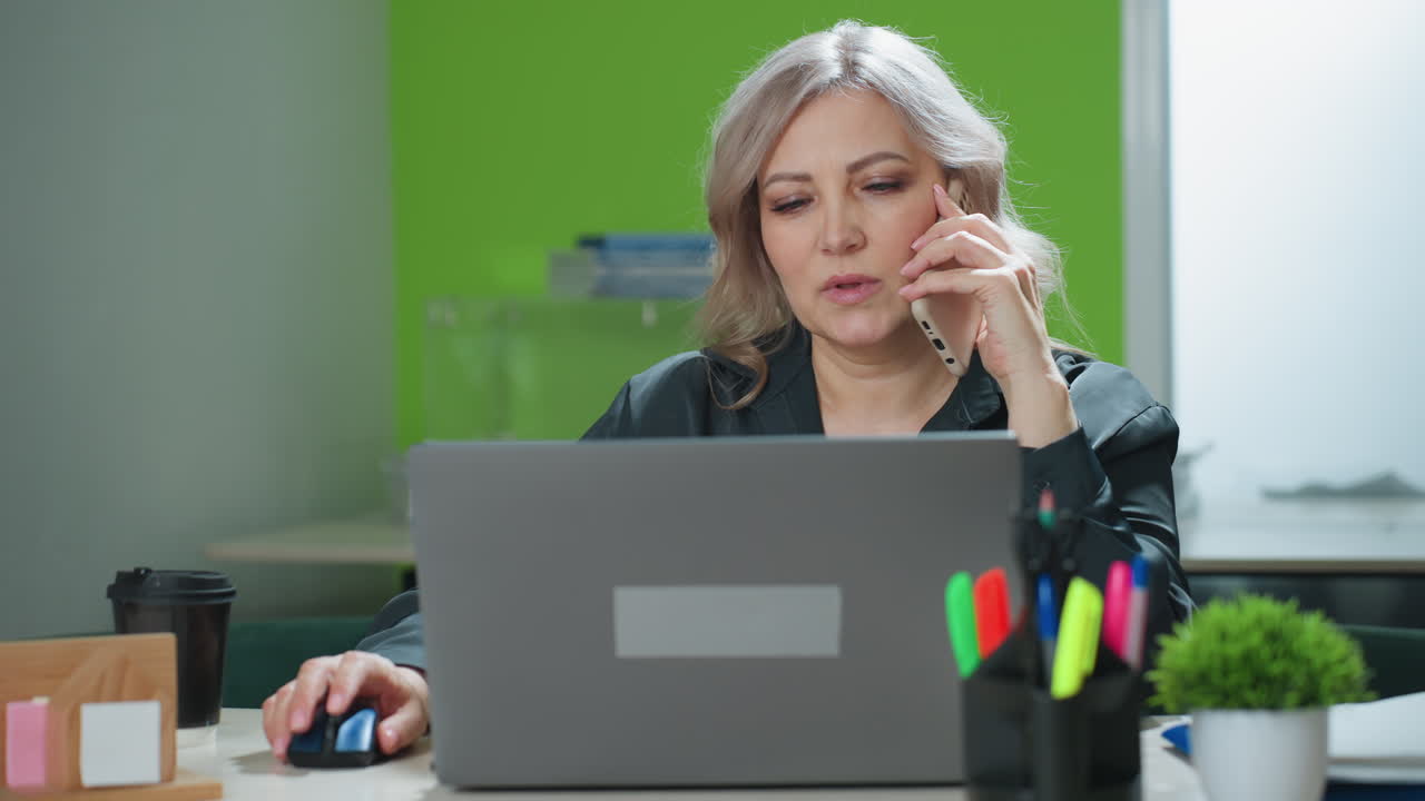 Confident real estate mogul sits at desk multitasking during phone conversation with client while viewing laptop screen in brightly lit office, focused on productivity and remote business discussion