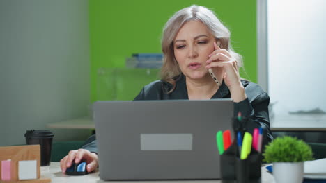 confident real estate mogul sits at desk multitasking during phone conversation with client while viewing laptop screen in brightly lit office, focused on productivity and remote business discussion