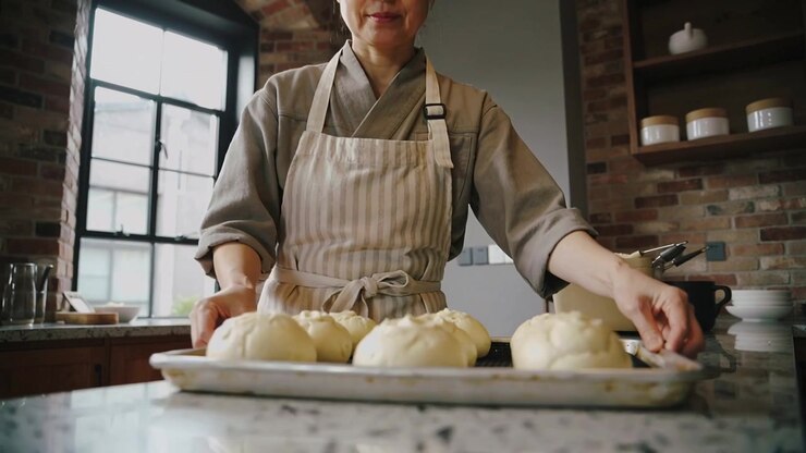 Picking up tray, baker nudging six buns on rustic kitchen counter for baking, wearing striped apron