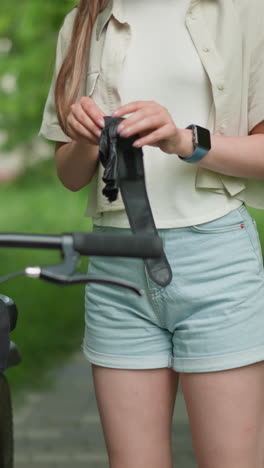 mujer joven en traje casual sostiene guantes negros, ponerlos cerca de la bicicleta estacionada en el camino pavimentado, ella usa reloj inteligente en la mano derecha con fondo borroso de vegetación y construcción